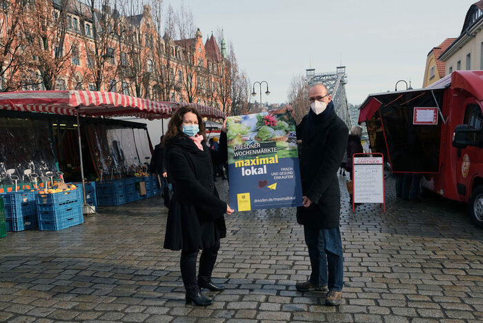 Mann und Frau halten ein Plakat, darum Marktstände, um Hintergrund das Blaue Wunder
