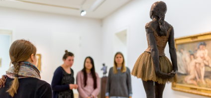 A woman with a plaited braid explains three other women's works of art. They are looking at the sculpture of a dancer.