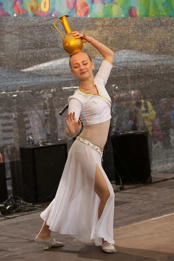 Eine Frau im weißen Gewand beim Tanzen mit einem goldenen Krug auf dem Kopf