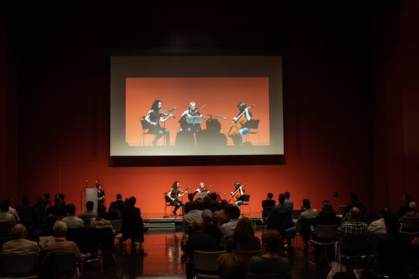 Das Publkium schaut Richtung Bühne und Leinwand, auf der drei Musiker*innen spielen.