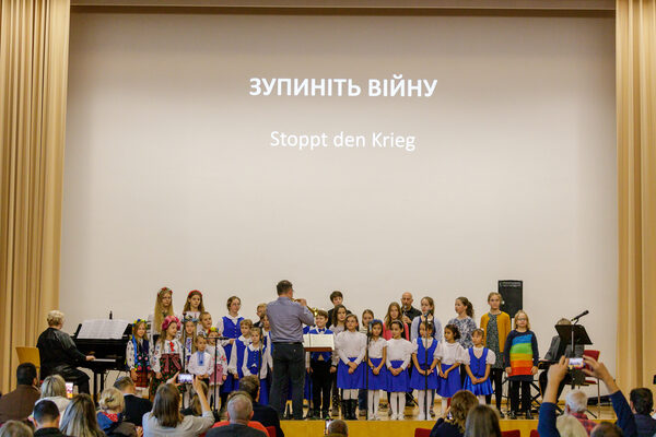 Kinder verschiedener Chöre stehen auf der Bühne und singen gemeinsam ein Lied. Im Hintergrund steht auf der Leinwand „Stoppt den Krieg“, als Liedtext geschrieben.