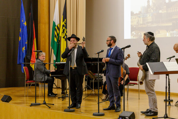 Drei Männer stehen jeweils mit einem Schofar-Horn auf der Bühne. Einer davon bläst in das Horn. Im Hintergrund sieht man zwei Musiker*innen, die am Klavier und mit einem Streichinstrument begleiten.