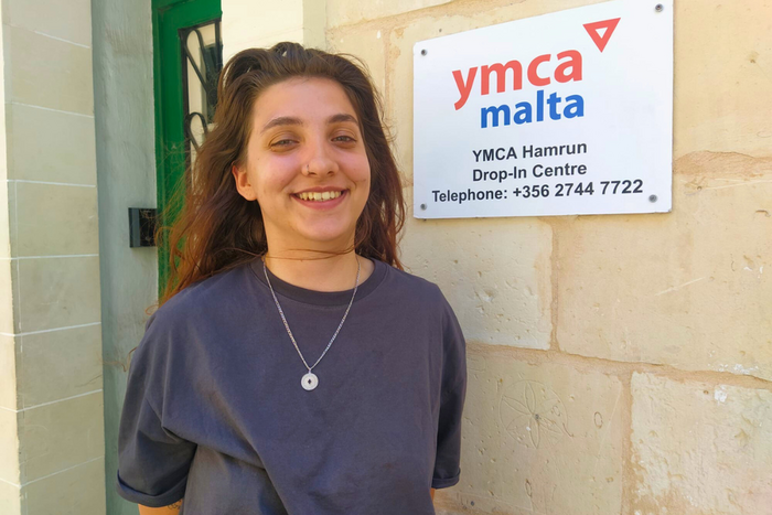 Junge Frau steht lächelnd vor einem Gebäude, an dessen Fassade ein Schild mit der Aufschrift YMCA Malta angebracht ist.