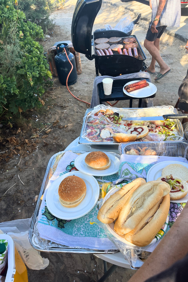 Verschiedene Burger, Hotdogs und Grillzutaten auf Campingtischen, im Hintergrund ein gut gefüllter Grill.