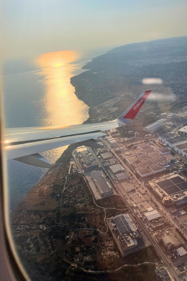 Blick aus dem Flugzeugfenster auf den Flügel, darunter Landschaft mit Gebäuden und Meer im Licht eines Sonnenuntergangs.