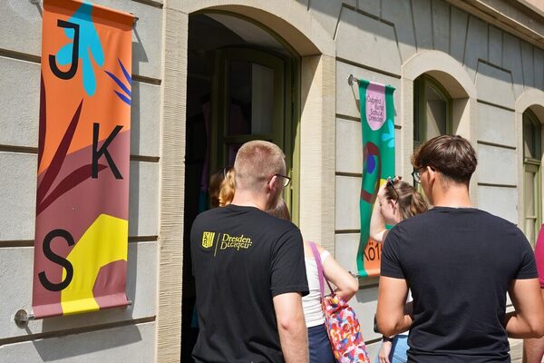 Junge Menschen mit Dresden-T-Shirt laufen in ein Gebäude, an dem bunte Banner mit der Aufschrift "JKS" hängen