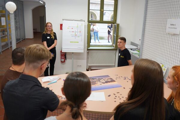 Eine kleine Gruppe junger Menschen steht mit dem Rücken zur Kamera und blickt auf ein Flipchart, auf dem ein Kreuzworträtsel zu sehen ist.