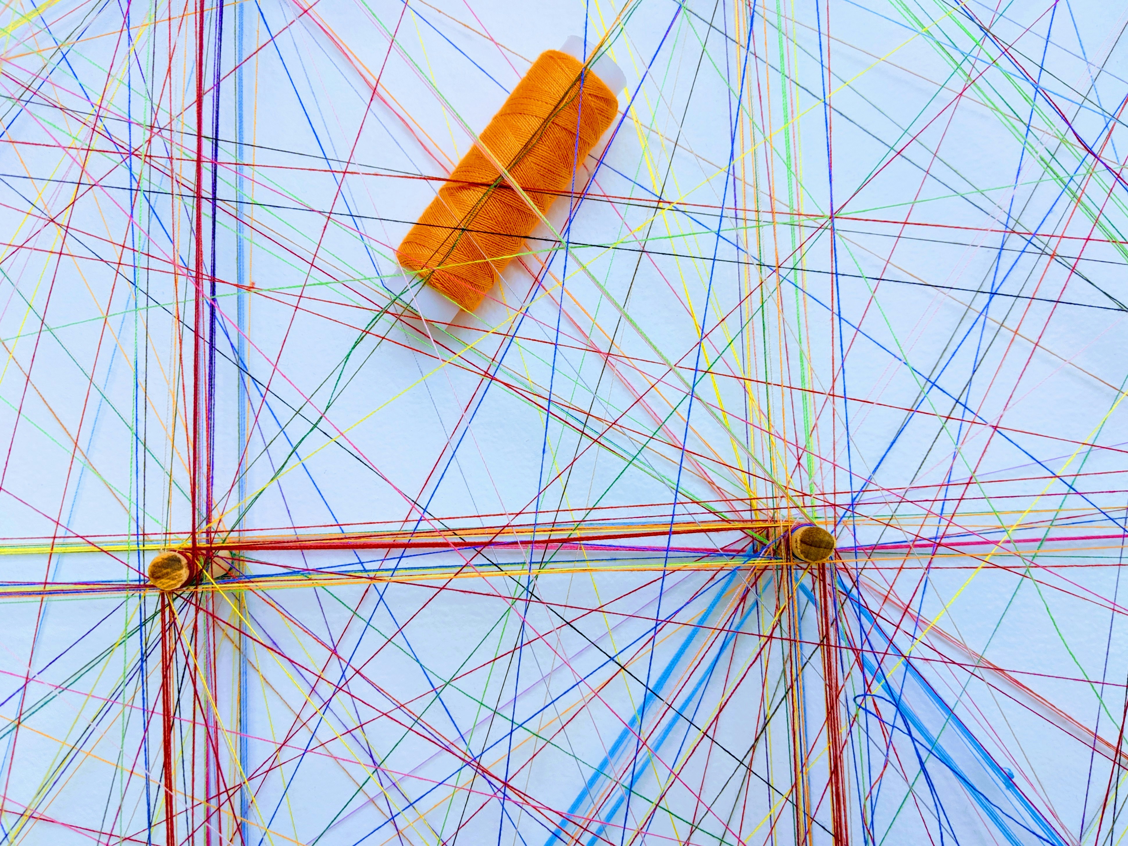 Ein Netz aus verschiedenfarbigen Bindfäden, eine Garnrolle mit orangefarbenem Bindfaden liegt darin
