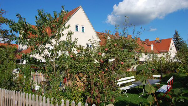 Blauer Himmel, Sonnenschein, nur eine einzige Wolke am Himmel. Das Bild gewährt einen Blick über den Zaun in eine Gartenidylle: Zu sehen ist eine Sitzgruppe mit historischen Gartenmöbeln, umrahmt von Apfelbäumen und Sträuchern, im Hintergrund eine Tessenowsche Häuserzeile.