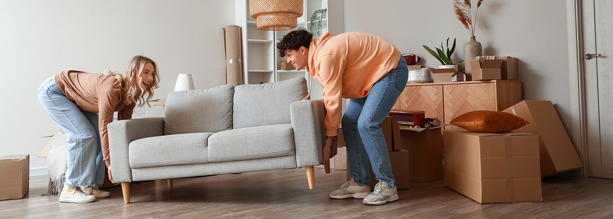 Eine junge Frau und ein junger Mann tragen ein graues Sofa in ihre neue Wohnung. Im Hintergrund stehen Umzugskartons