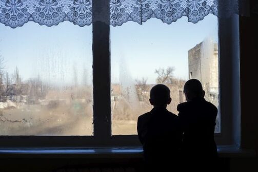 zwei Kinder schauen durch das Zimmerfenster in die zerstörte Umgebung