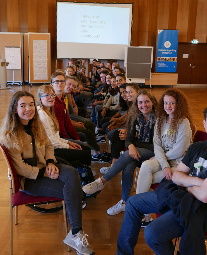 Schülerinnen und Schüler sitzen sich in zwei Reihen gegenüber. Im Hintergrund befinden Sie eine Leinwand und Stellwände.