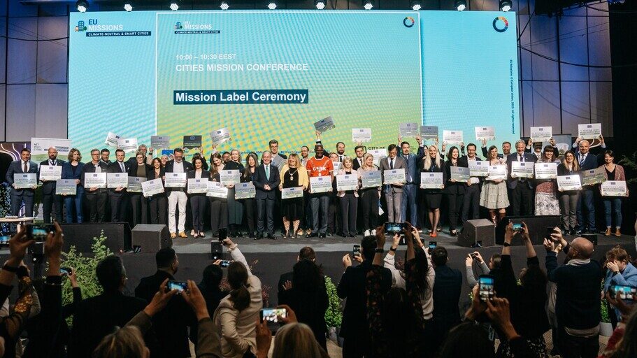 Eine Gruppe Menschen steht auf einer Bühne vor einer Leinwand mit der Schrift "Mission Label Ceremony"