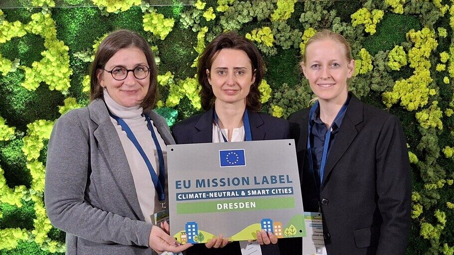 Gruppenfoto der Dresdner Delegation bei der MissionCities-Konferenz 2025 in Vilnius: Dr. Franziska Graube-Kühne, Radka Roßner und Regine Kramer (v. l.) stehen vor einer grünen Wand im Foyer. Regine Kramer hält das Mission-Label in den Händen.