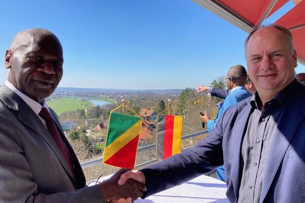 Zwei Männer schütteln sich die Hände, dahinter auf einem Tisch eine kongolesische und eine deutsche Flagge