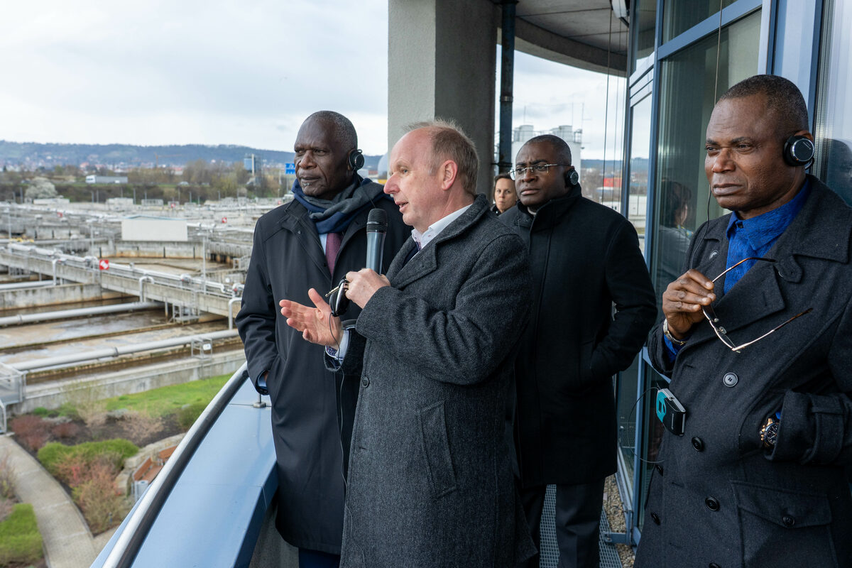Ein Mann erklärt drei Männern mit Kopfhörern etwas, im Hintergrund eine Kläranlage