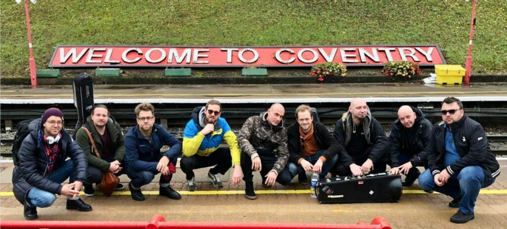 Die Band Yellow Cap in Coventry: Die neun Musiker hocken vor einem Rasenstück und einem Banner mit der Aufschrift "Welcome to Coventry"