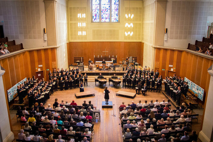 Blick in den Kirchensaal: Recht und links mehrere Reihen von Chorsängern, in der Mitte zwei Flügel und voll besetzte Publikumsreihen