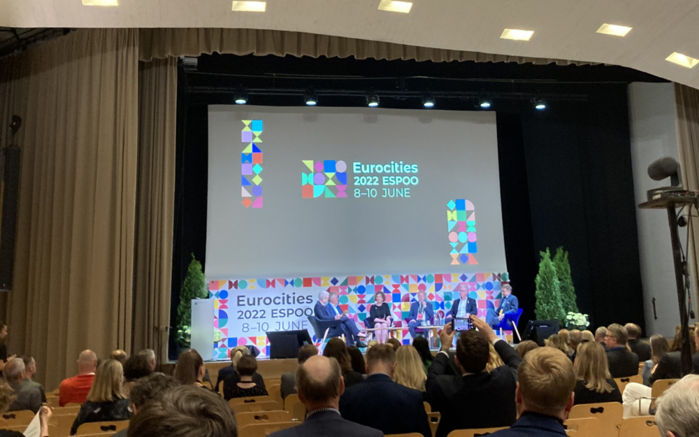 Blick über das Publikum auf die Bühne und eine Paneldiskussion, im Hintergrund das Logo der Veranstaltung