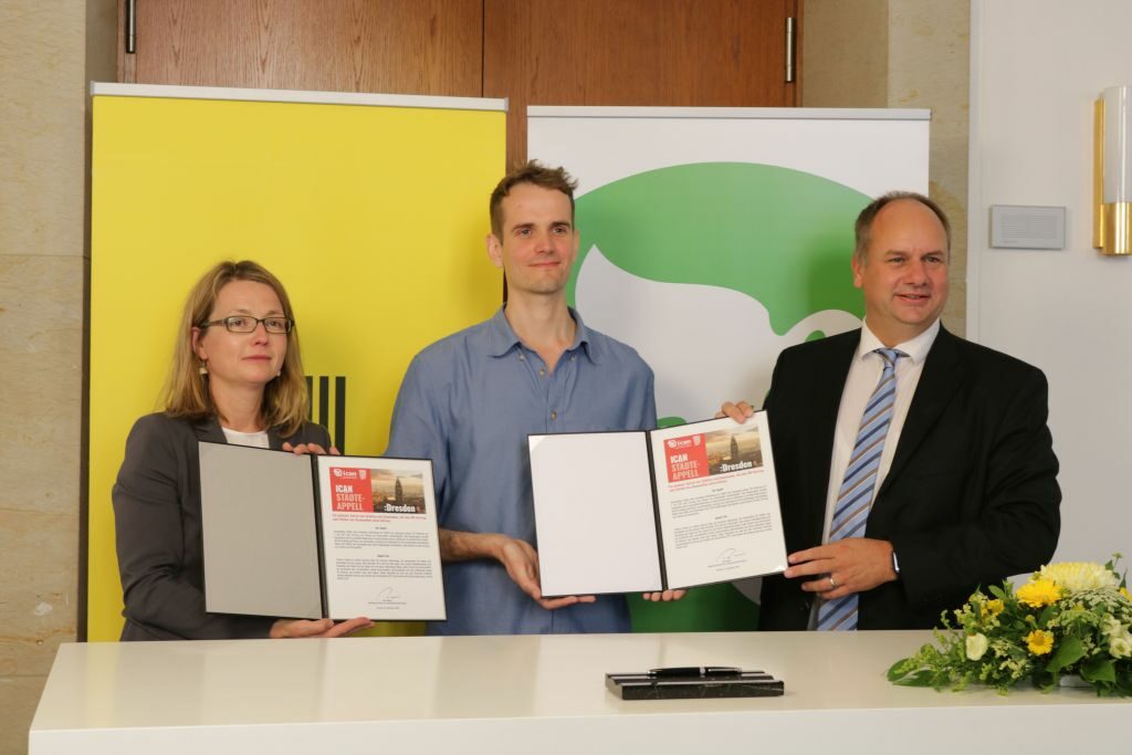 OB Dirk Hilbert, die Zweite Bürgermeisterin Annekatrin Klepsch und ICAN-Botschafter Emanuel Wenzel stehen mit der unterzeichneten Urkunde des ICAN-Städteappells am Unterzeichnungstisch