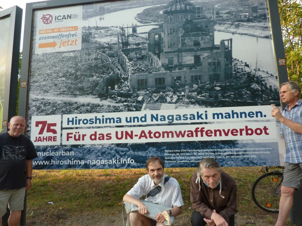 Akteure der Mahnwache vor einem großen Plakat mit dem Motiv von Hiroshima nach dem Atombombenabwurf und der Aufschrift: "Hiroshima und Nagasaki mahnen. Für das UN-Atomwaffenverbot"