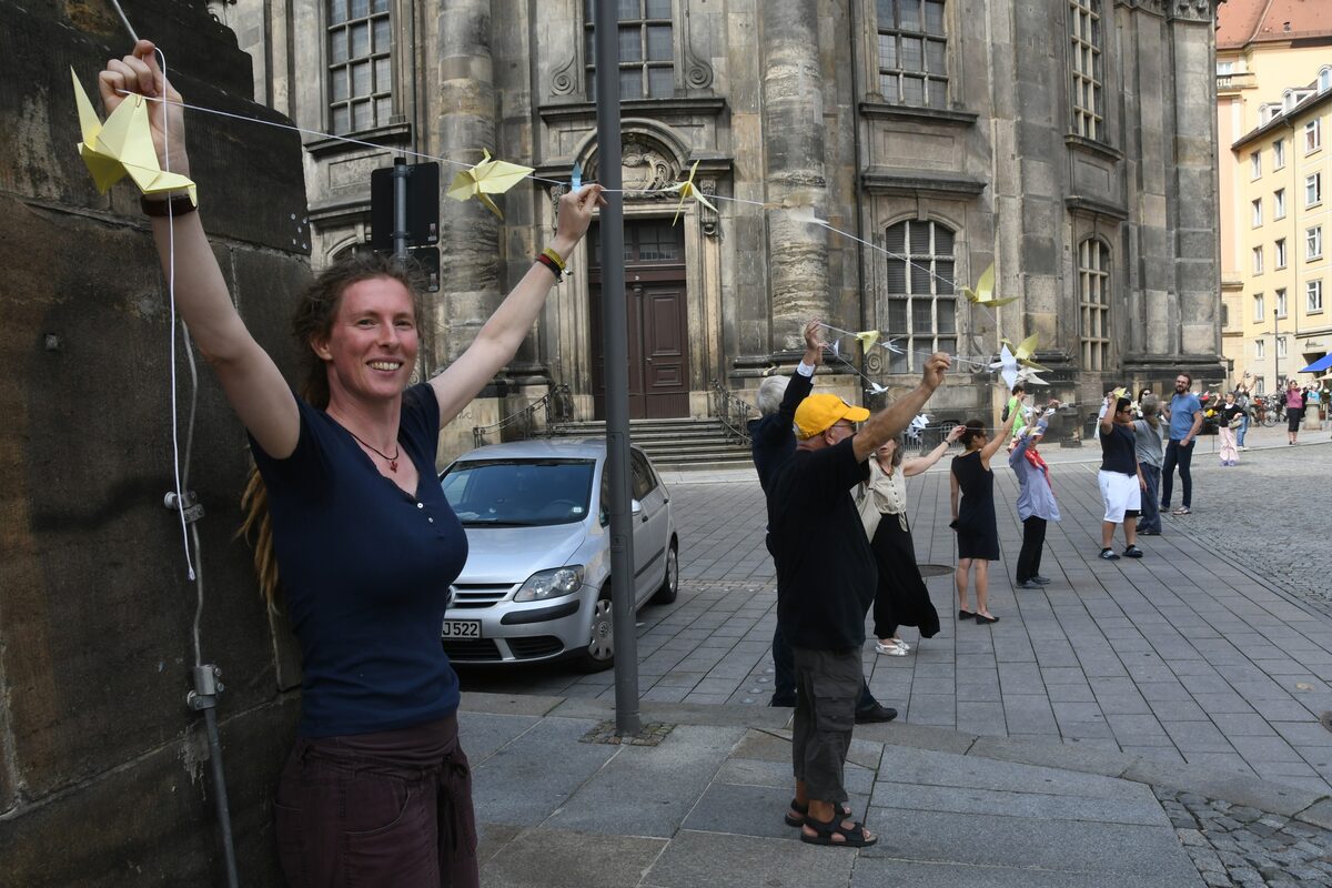Menschenkette mit Origami-Kranichen vor der Kreuzkirch über die Kreuzstraße, Menschen heben die Kette mit den Kranichen hoch als Symbol: Lasst sie fliegen!