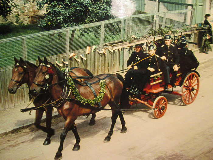 historische Aufnahme: Feuerwehrleute auf einem von Pferden gezogenen Wagen