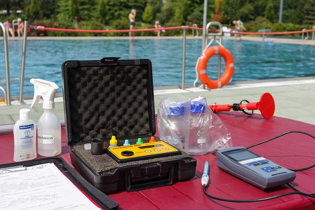 Auf einem Tisch im Vordergrund befinden sich Instrumente und Geräte zur Bestimmung der Wasserqualität. Im Hintergrund ist ein Schwimmbecken.