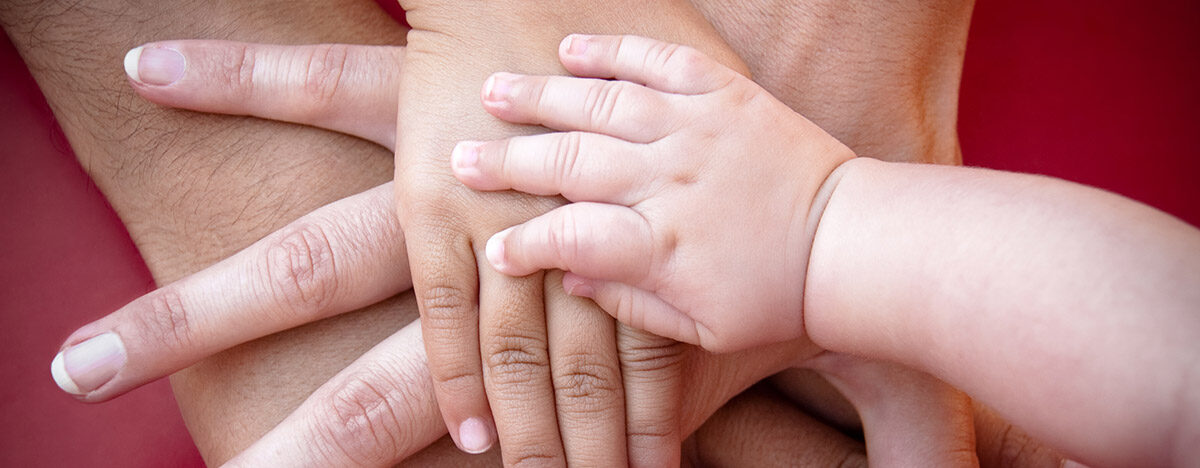 vier aufeinander gelegte Hände von unterschiedlichen Personen, die oberste ist die Hand eines Kleinkindes