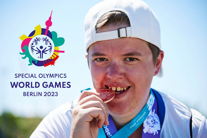 Ein junger Mann testet seine gewonnene Medaillie auf Bissfestigkeit. Zusätzlich ist das Logo der Special Olympics World Games 2023 zu sehen.