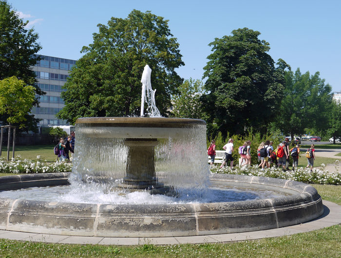 Springbrunnen im Blüherpark mit Sandsteinbecken und oberer Schale mit Fontäne, im Hintergrund Bäume und Menschen