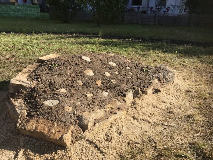 in der Rasenfläche befindet sich eine erhöhte Fläche aus Sand-Erde-Gemisch, umrandet mit Steinen