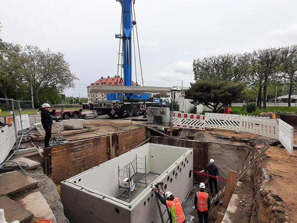 Blick in die Baugrube, der Technikschacht aus Betonfertigteilen wurde eingehoben, Kran und andere Baumaschinen sowie Bauleute im Einsatz