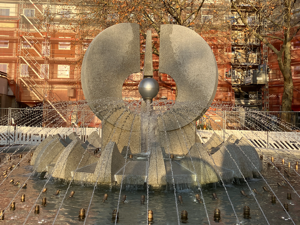 Krachtbrunnen: kreisrunde Brunnenschale, mittig die Brunnenfigur mit Metallkugel; das Wasser kommt aus den kreisförmig angeordneten Düsen