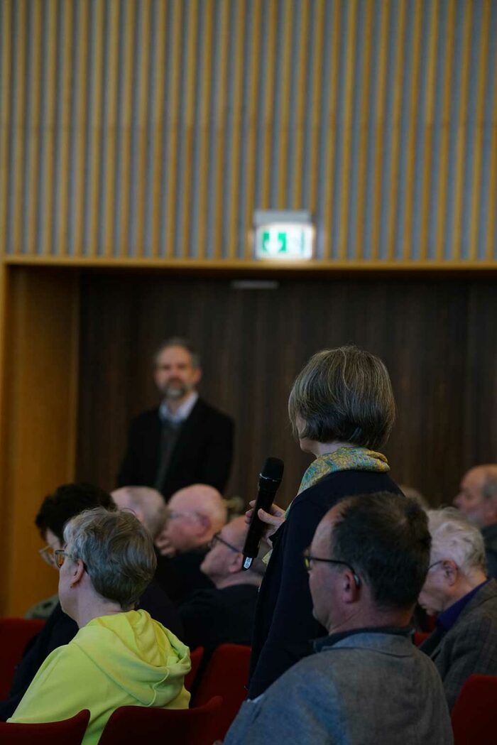 Eine Teilnehmerin spricht in ein Mikrofon, weitere Teilnehmer sitzen im Saal