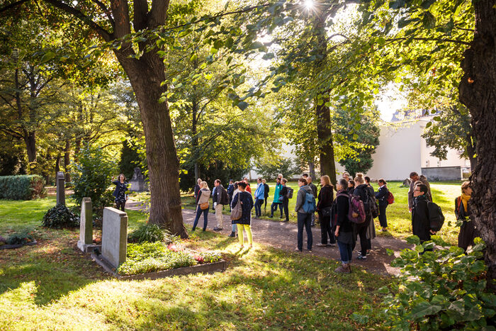 Menschen besichtigen den Neuen Annenfriedhof. Sie stehen vor in einer Wiese befindlichen Gräbern. Rundherum befinden sich alte Lindenbäume.