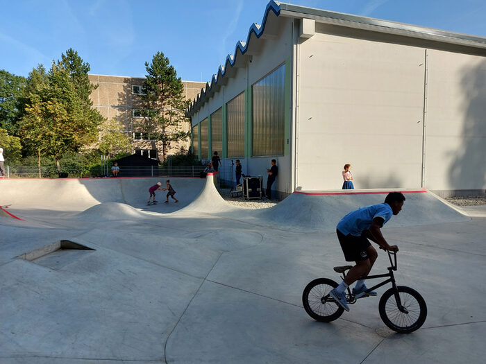 Skateanlage mit verschiedenen Elementen, im Vordergrund ein Sportler mit Bike, im Hintergrund die benachbarte Turnhalle