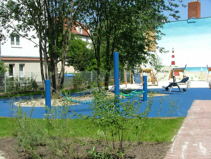 Spielplatz "Löbtauer Strand" am alten Standort Reisewitzer Straße 73. Im Vordergrund Hängematten auf blauem Fallschutz, der das WAsser darstellt. Im hintergrund weitere Spielgeräte. Diese stehen vor einer mit einer Strandlandschaft bemalten Hauswand.