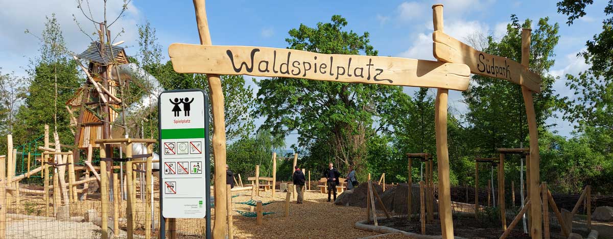 Eingangschild zum Waldspielplatz, im Hintergrund Spielgeräte aus Holz