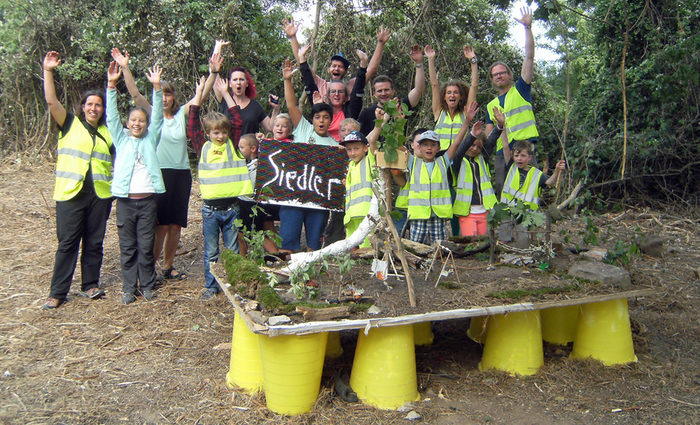 Gruppe Kinder und Erwachsener mit erhobenen Händen hinter einem Modell mit einem gestalteten Waldspielplatz