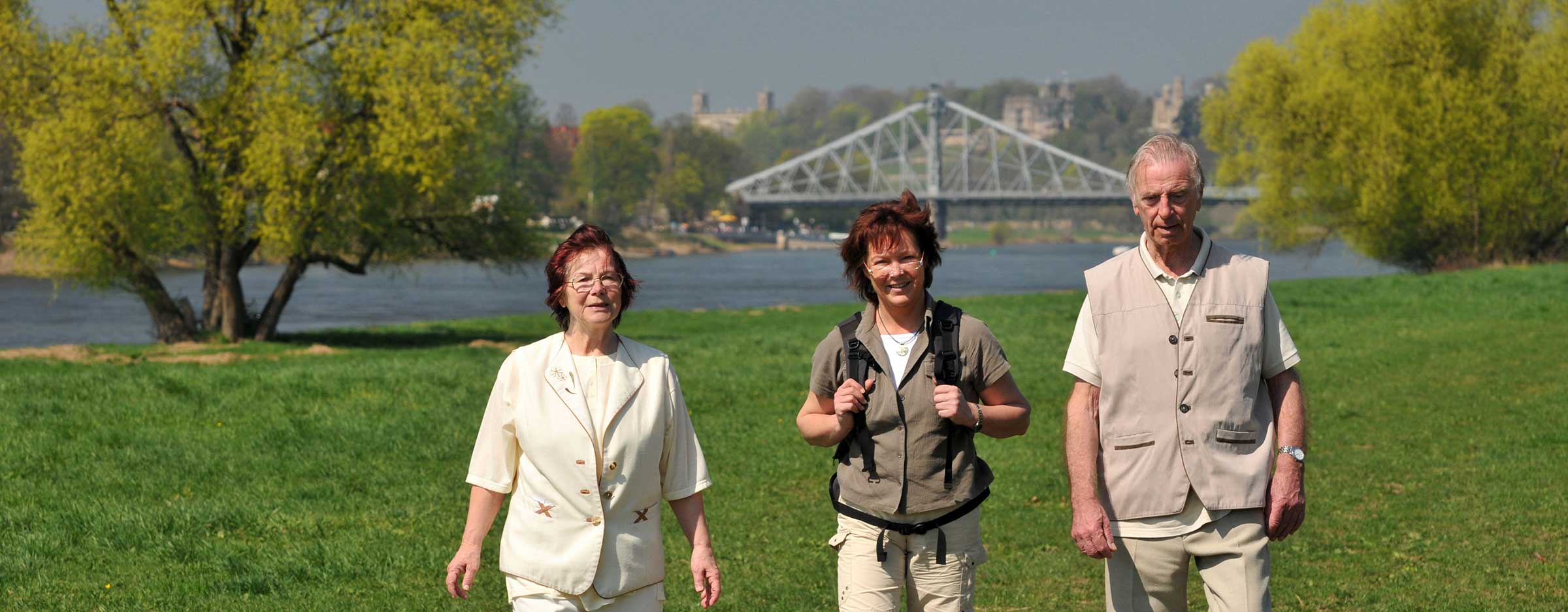Zwei ältere Frauen und ein älterer Mann stehen auf der Wiese vor einem Fluss, im Hintergrund die Brücke Blaues Wunder