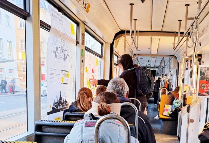 Mehrere Menschen sitzen in einer Straßenbahn. Eine Person klebt einen Notizzettel an ein Plakat.