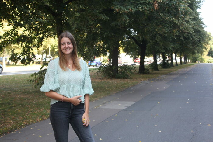 Eine Frau steht auf der Straße, hinter ihr befindet sich eine grüne Baumallee