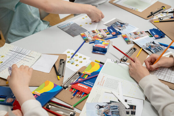 Kinderhände an einem Tisch mit Klemmbrettern, Buntstiften und Stickern. Die Kinder gestalten ein Architekturleporello aus.