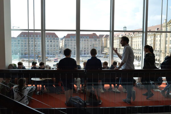 Die Schülerinnen und Schüler sitzen oder stehen im Kulturpalast Dresden an der Fensterfront mit Blick auf den Altmarkt. Ihnen wird die Innengestaltung des Kulturpalast beschrieben.