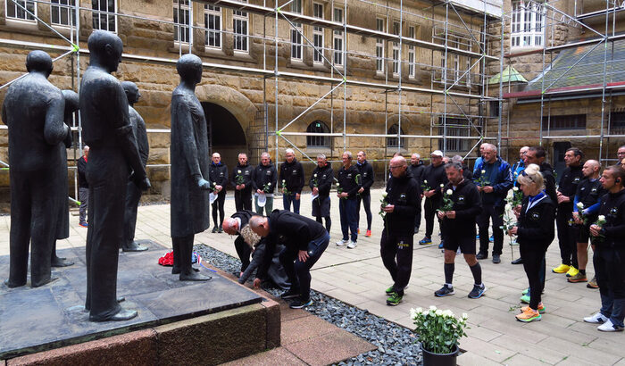 Sportlerinnen und Sportler legen weiße Rose vor einem Denkmal nieder.