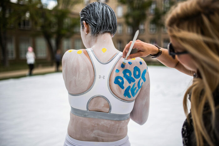Im Rahmen einer Performance wird einer Frau, die am ganzen Körper mit weißer Farbe bemalt ist, das Wort Protect in blauer Farbe auf den Rücken gemalt.