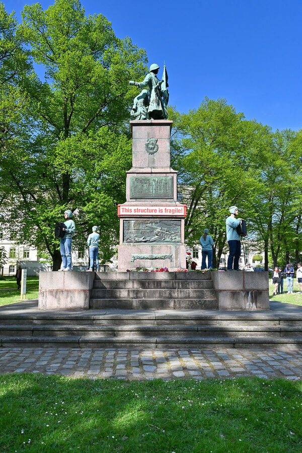 Denkmal aus Granit mit Bronzereliefs am Sockel zeigt Bilder des Kampfgeschehens. Vier Menschen stehen auf dem Sockel für eine Performance.