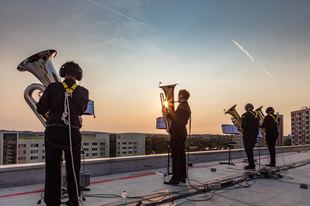 Blasmusiker stehen auf dem Dach eines Plattenbaus in Prohlis, im Hintergrund der Himmel und weitere Plattenbauten und Hochhäuser