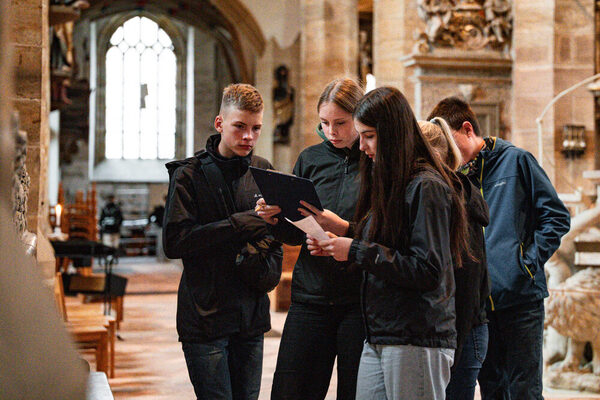 Vier Personen stehen in einer Kirche, zwei halten ein Klemmbrett, alle tragen dunkle Jacken.
